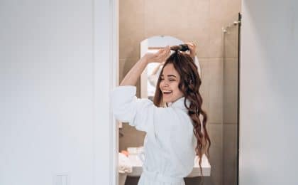 Mulher alegre em roupão fazendo cachos no cabelo com um babyliss como parte de sua rotina de autocuidado matinal.