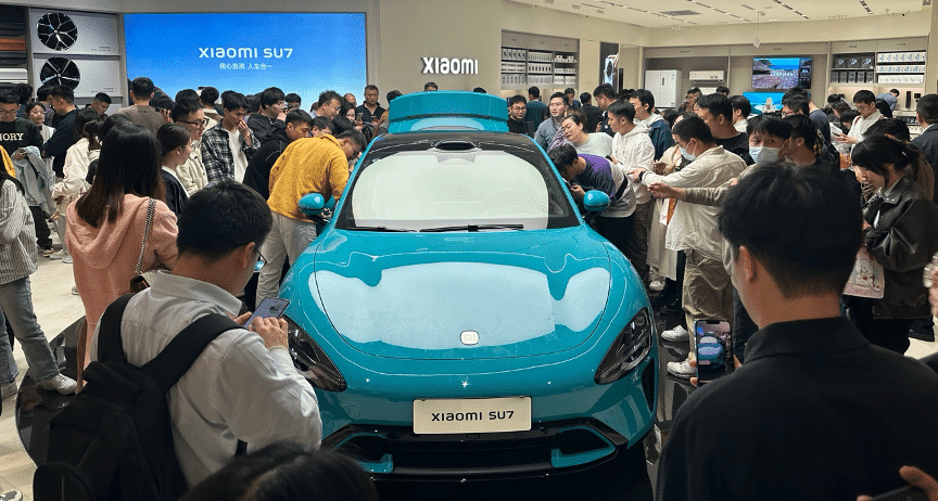 Público reunido ao redor de um veículo Xiaomi SU7 azul em uma loja de experiência da Xiaomi em Pequim, mostrando grande interesse e examinando o carro de perto.